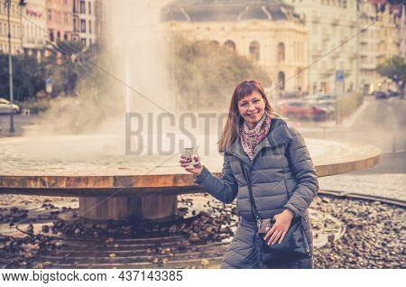 Young Woman Tourist With Grey Jacket Smile And Hold Therapeutic Mineral Water In Cup Near Fountain H