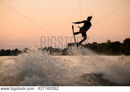 Active Guy Jumping Over Splashing Wave On Wakeboard Holding On To The Rope. Water Sports Activity.