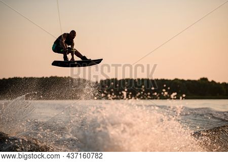 Active Muscular Man Jumping High With Wakeboard Over Splashing Water Against The Backdrop Of Sky