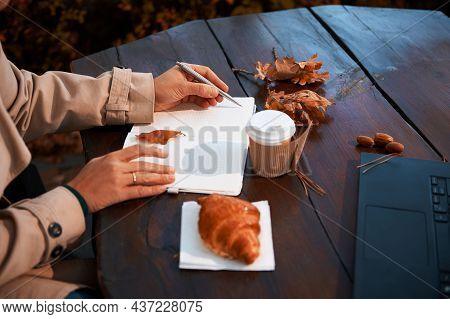 Focus On Female Hand Holding Silver Pen And Writing The List To Do In Notepad, While Sitting At A Wo