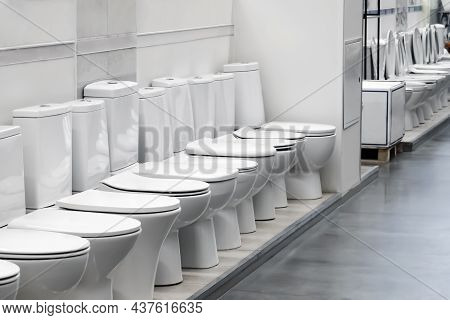 Exhibition Of Samples Of White Toilet Bowls Standing In A Row In The Warehouse Of A Plumbing Store. 