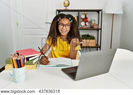 Young african american girl doing homework at home smiling with happy face looking and pointing to the side with thumb up. 