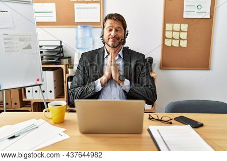 Handsome middle age man wearing call center agent headset at the office praying with hands together asking for forgiveness smiling confident. 