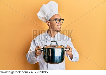 Bald man with beard wearing professional cook holding cooking pot smiling looking to the side and staring away thinking. 