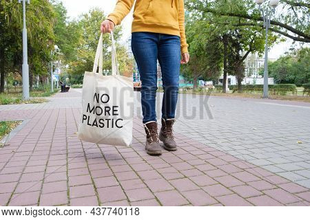 A Woman Walks Down The Street And Holds A Reusable Cloth Bag That Says No More Plastic.