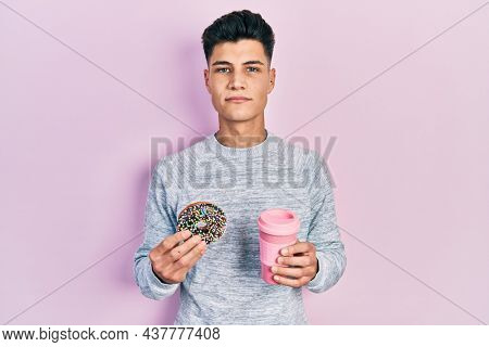 Young hispanic man eating doughnut and drinking coffee relaxed with serious expression on face. simple and natural looking at the camera. 