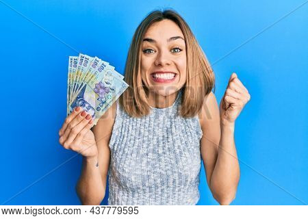 Young caucasian blonde woman holding 100 romanian leu banknotes screaming proud, celebrating victory and success very excited with raised arm 