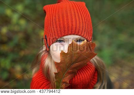 Portrait Of A Girl In An Orange Hat With An Autumn Leaf Against The Background Of The Forest