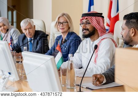 Group of international politicians sitting at table with national flags and listening to speech of Islamic representative in ghutrah at conference