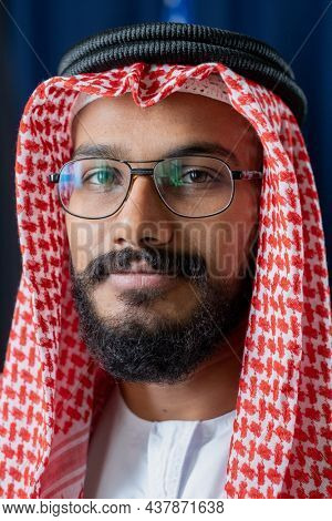 Content Islamic man with beard wearing glasses, agal and ghutrah looking at camera against dark blue curtains
