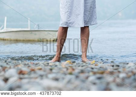 A Girl Walks Along A Rocky Shore With Bare Feet, Her Feet In The Water