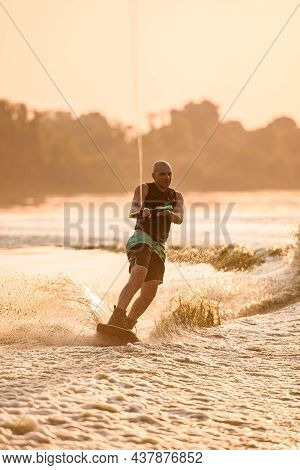 Energy Strong Man Engaged In Water Sport And Holds Rope And Speedly Riding Wakeboard On The River.