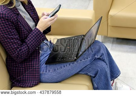 Girl In Jeans And Checkered Jacket Sitting On Leather Sofa With Smartphone And Laptop On Her Knees. 