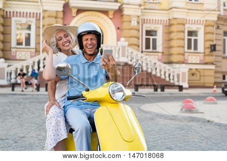Couple on scooter with phone. Man and woman smiling widely. Send a video message. We're having a great time.