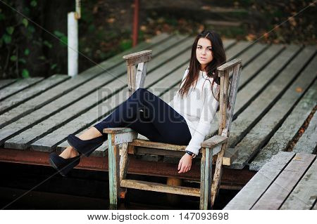 Beautiful brown-eyed brunette with long hair tousled by the wind, a white blouse, dark blue leggings and trendy shoes on a high platform, sitting on wooden stairs at the pier over the water.