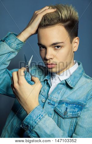 Portrait of fashion blonde Barber hairstylist with scissors in hand near eye on blue background.