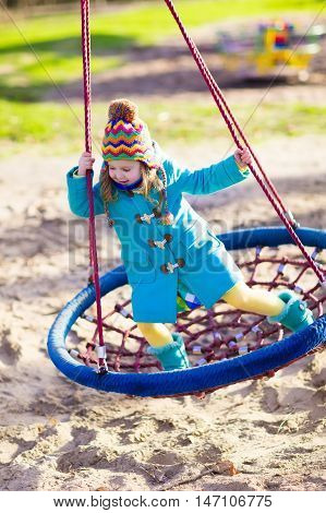 Little girl in warm coat and hat on a playground. Child playing outdoors in autumn. Kids play on school yard. Happy kid in kindergarten or preschool. Children having fun on cold fall day. Toddler on a swing.
