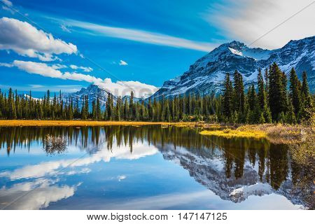 Snow-covered mountains are reflected in smooth water of the lake. Rocky Mountains on a sunny autumn day. The concept of active tourism and ecotourism