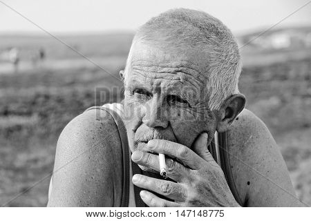 An elderly man smokes in nature. He is thoughtful and lonely.