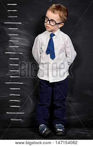 Four year old boy in white shirt and a tie standing by a stadiometer, drawn on a blackboard. Education.