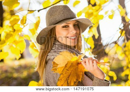 Beautiful young woman in park in autumn holding leaves. Closeup outdoor portrait of happy cheerful young Caucasian woman in gray sweater, floppy hat and scarf posing. Vibrant colors, natural light.