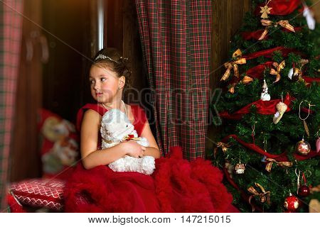 Little winter Princess - girl in red dress sitting on windowsill looks out window at night. Celebration of New year and Christmas in enchanting holiday interior with decorated pine