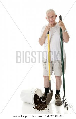 A senior man looking at the viewer as he holds a garden rake and shovel, a bucket of spilled dirt at his feet.  On a white background with space on his right for your text.