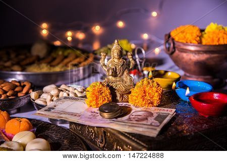 oil lamp or diya with crackers, sweet or mithai, dry fruits, indian currency notes, marigold flower and statue of Goddess Laxmi on diwali night while laxmi pujan or pooja