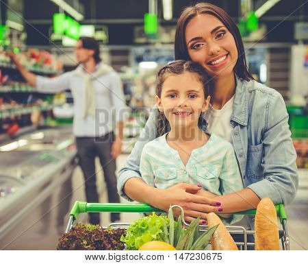 Family in the supermarket. Beautiful young mom and her little daughter are looking at camera and smiling in the background dad is choosing goods