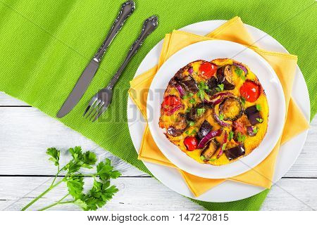 oven baked tasty omelet with eggplantred onion and tomato sprinkled with parsley on white dish with fork and knife on table mat top view