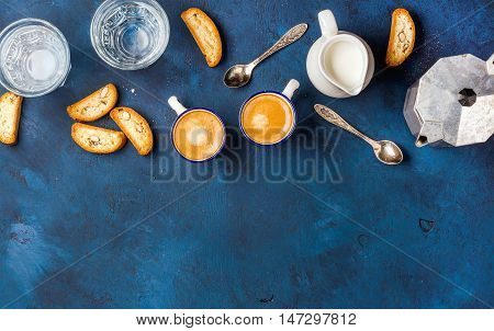 Coffee espresso in cups with italian cantucci, cookies and milk in jug over dark blue painted plywood background, top view, copy space, . Food frame concept