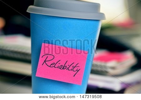 closeup of a pink sticky note with the word reliability in a blue cup of coffee or tea on an office desk