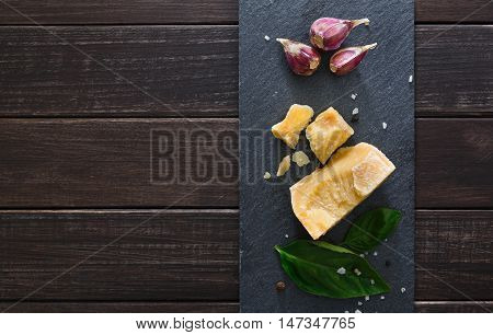 Cheese delikatessen closeup on black stone desk at wooden surface background. Parmesan pieces decorated with basil and garlic, top view image with copy space