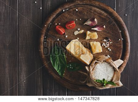 Cheese delikatessen closeup on rustic wood. Wooden desk with parmesan, camembert and brie cuts decorated with garlic, tomato and basil, top view image with copy space