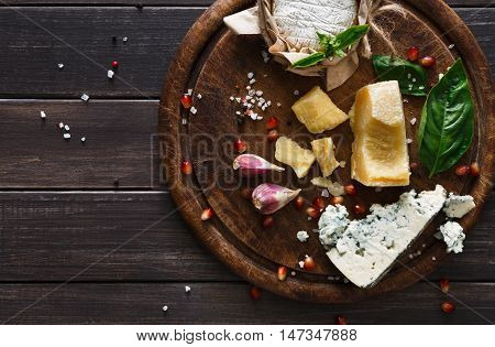 Cheese delikatessen top view on rustic wood. Wooden round desk with blue cheese roquefort, parmesan, camembert and brie cuts decorated with garlic, pomegranate and basil, french cuisine ingredients