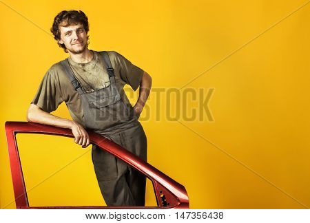 Handsome smiling mechanic posing in work clothes, he leans on the red car's door on the background of yellow bright wall