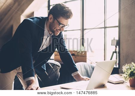 Best solutions every day. Confident young man in smart casual wear looking at laptop and smiling while standing near his working place in office