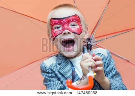 Excited caucasian boy with a spiderman mask on his face smiles at camera while holding an umbrella.