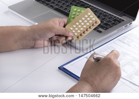 doctor hand holding birth control pills and pen over computer and patient history form on white table healthcare pharmacy and medicine concept