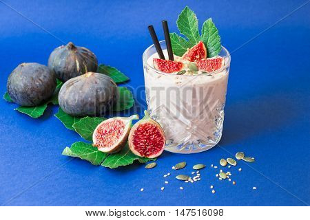 Glass of fig fruit smoothie fig slices straws and near scattered sunflower susami seeds ripe fig fruits on the fig tree leaves on blue background. Fig fruit smoothie glass. Horizontal. Daylight.