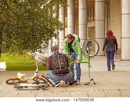 MOSCOW, RUSSIA - September 4, 2016: Man in the squatting position with a backpack with two umbrellas corrects the malfunction of an inverted bicycle in the park Museon. Standing mid adult woman watching his actions. September 4, 2016 in Moscow, Russia