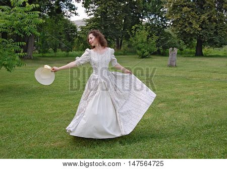 European woman dancing and touching her hat in vintage dress in park. White color, curly hair, sensitive, sensuality.