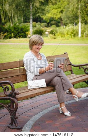Attractive mature woman sitting on a bench holding and using a digital tablet in the park at summer day