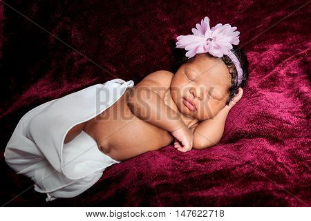Sweetly sleeping African American newborn girl. She is loosely wrapped and posed on her side with hand under her face. She is laying on a maroon blanket.