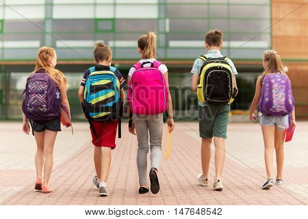 primary education, friendship, childhood and people concept - group of happy elementary school students with backpacks walking outdoors from back