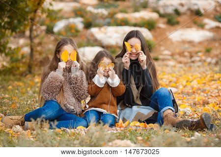 Three girls, three sisters eastern appearance, older, middle and younger, one brunette and two blonde girls, all three dressed in blue jeans and brown shoes, playing together in a beautiful autumn park with crumbling yellow and red leaves