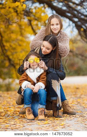 Three girls, three sisters eastern appearance, older, middle and younger, one brunette and two blonde girls, all three dressed in blue jeans and brown shoes, walking together in a beautiful autumn park with crumbling yellow and red leaves