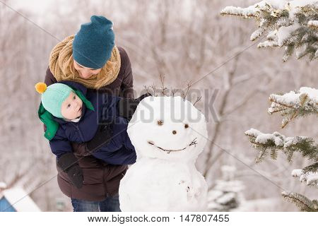 Portrait of happy mother with cute toddler boy with funny snowman outdoors. Mom with adorable child walking in the winter park. Christmas and winter concept.