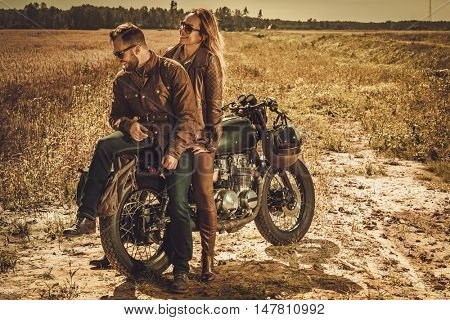 Young, stylish cafe racer couple on the vintage custom motorcycles in a field.