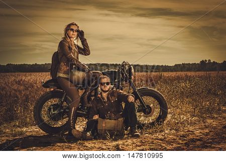 Young, stylish cafe racer couple on the vintage custom motorcycles in a field.